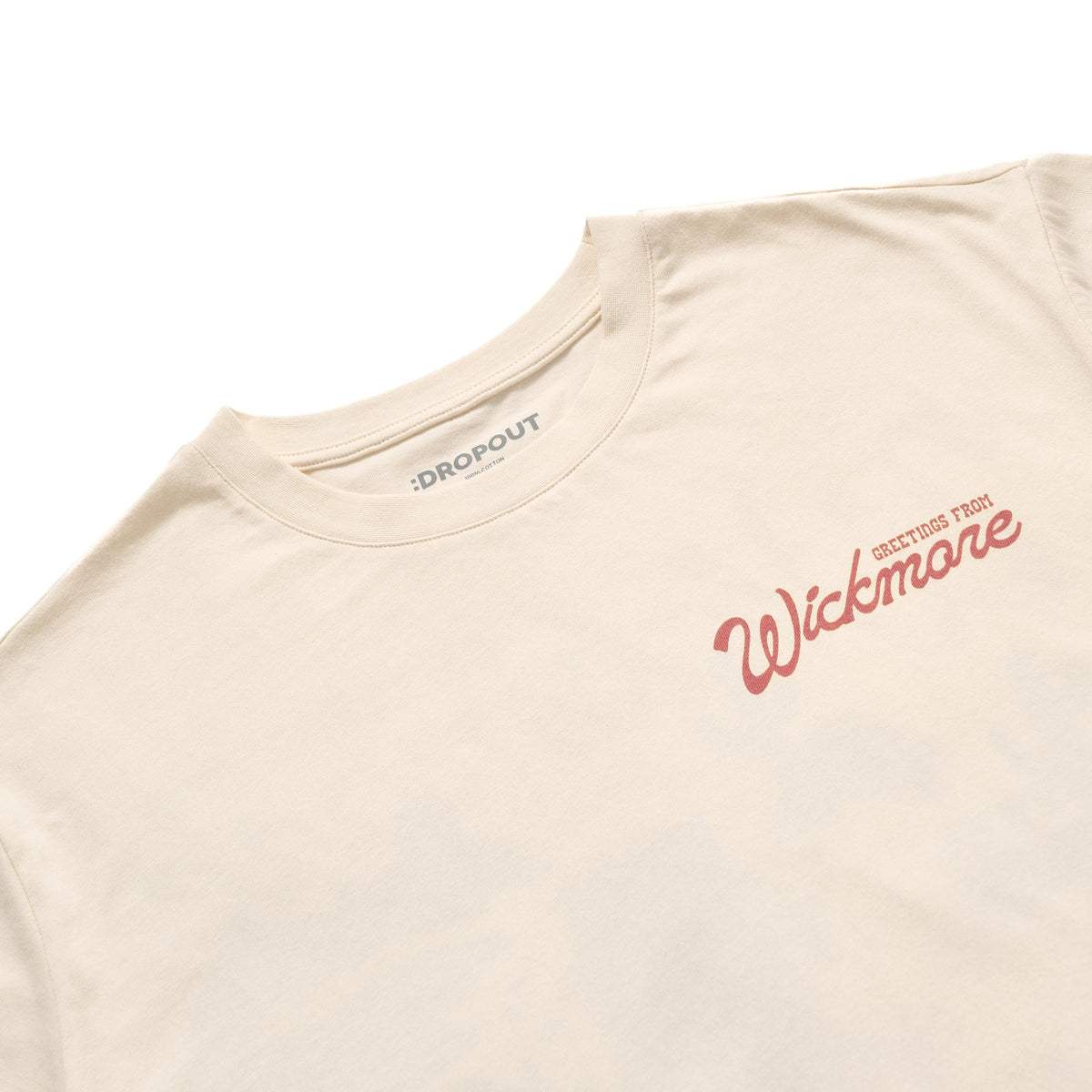 A close-up of a cream-colored t-shirt with a red script logo reading 'Wickmore' and smaller text 'GREETINGS FROM' on the chest, with the brand 'DROPOUT' visible inside the collar.