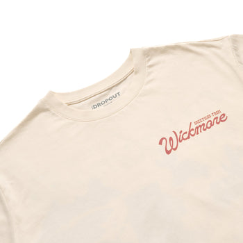 A close-up of a cream-colored t-shirt with a red script logo reading 'Wickmore' and smaller text 'GREETINGS FROM' on the chest, with the brand 'DROPOUT' visible inside the collar.
