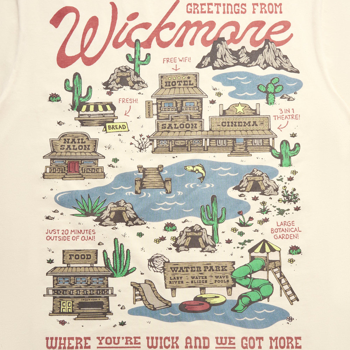 A close-up of a cream-colored t-shirt featuring a detailed western-themed graphic print that reads 'Greetings from Wickmore'. The illustration includes cacti, mountains, a saloon, a pool with a green slide, and whimsical text.