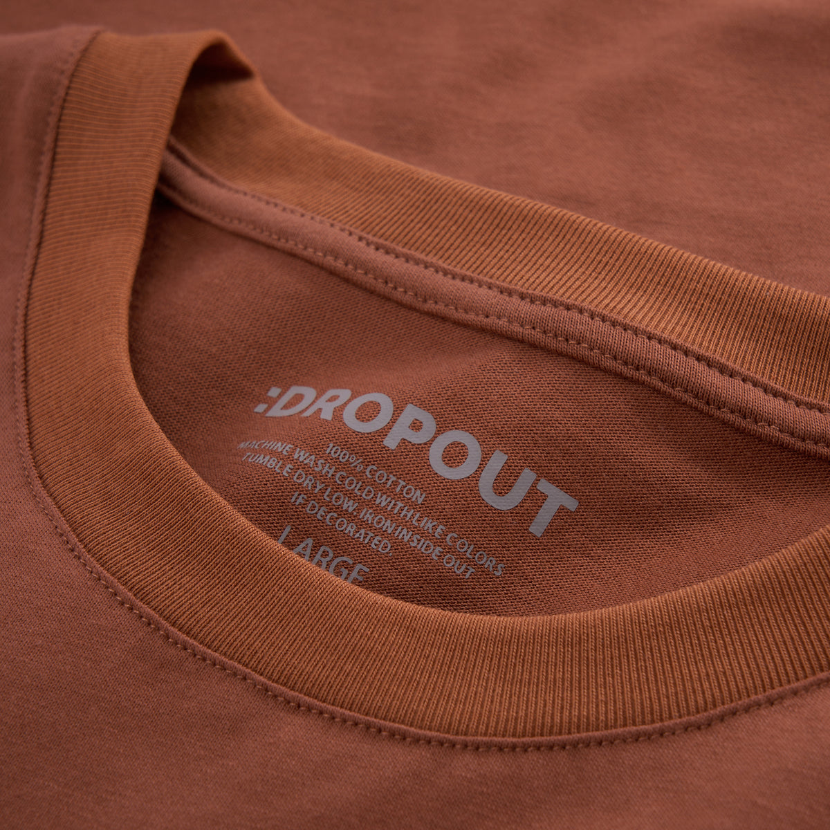 Close-up of a rust-brown crewneck t-shirt showing the inner collar print with the brand ':DROPOUT' and size 'LARGE'.